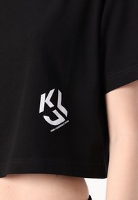 Svart croppet t-skjorte laget av bomull, med et hvitt grafisk logo på venstre bryst. Korte ermer med rund hals og ren hem.