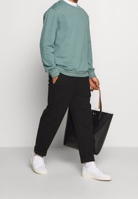 Hellgrüner Pullover, schwarze schmal zulaufende Hose und weiße Sneakers. Der Mann trägt eine schwarze Tragetasche mit einem beigen Riemen. Einfaches Design, lockere Passform.