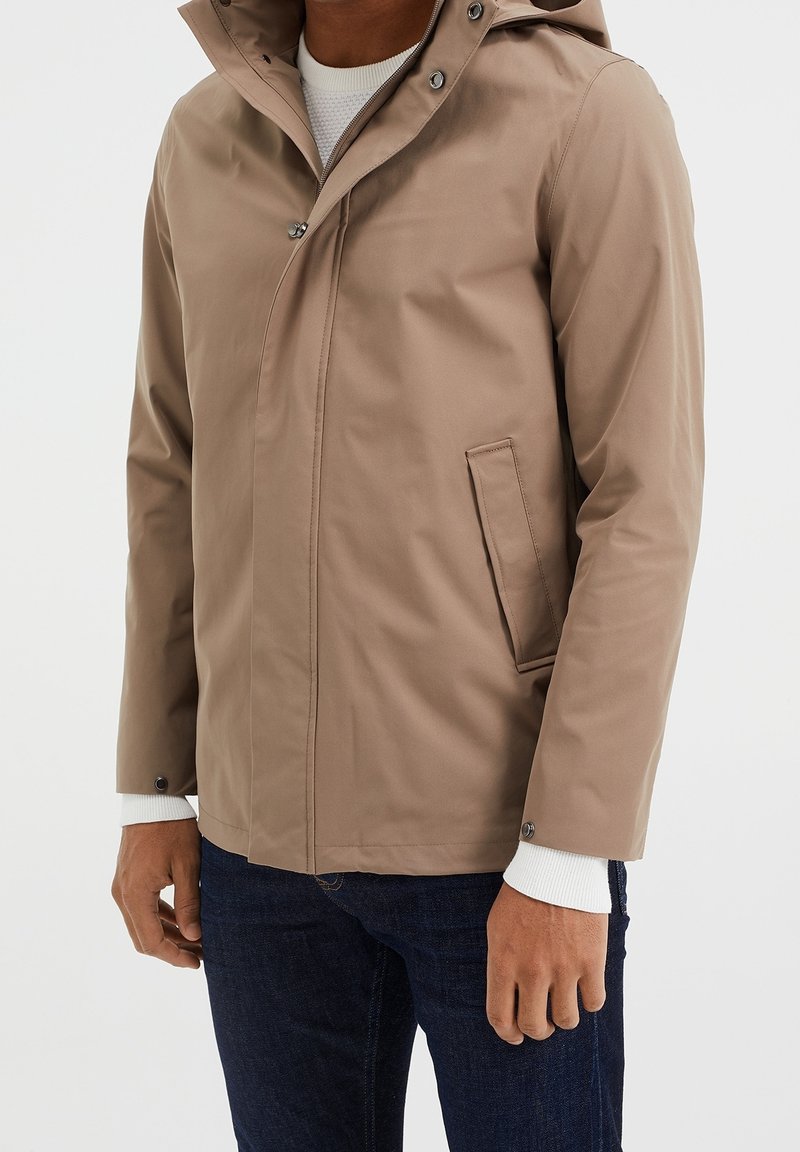 Giacca impermeabile marrone con cappuccio a punta, bottoni a pressione, tasche laterali e texture liscia. Indossata sopra un maglione bianco lavorato a maglia.