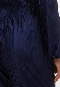 Lahtise vertikaalse triibumustriga tumesinine volditud kleit, mis on läikiva tekstuuriga, pikkade varrukatega ja keha ümbritseva vöökohaga.