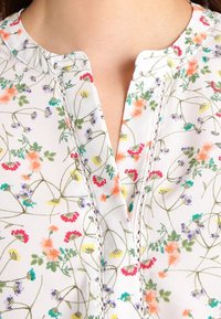 Kukoita blusa floral na kitambaa chepesi, cheupe. Ina shingo ya V yenye kushona kwa uangalifu, imepandwa na maua ya rangi mbalimbali na shina za kijani.