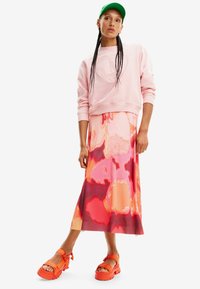 Sudadera rosa con logo en relieve, combinada con una falda maxi de colores rosa y naranja. Sandalias anchas naranjas. Gorra verde opcional.