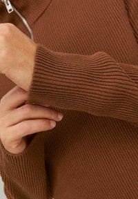 Suéter marrón acanalado, con un cuello con cremallera. La textura es densa con líneas verticales, y los puños de las mangas son ligeramente holgados.