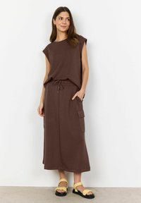 Brown sleeveless top paired with a long brown skirt featuring side pockets. Model wears yellow sandals with ruffled straps.