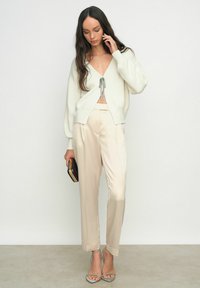 Bílý pletený kardigan s hlubokým výstřihem do V a detailem na zavazování, kombinovaný se světle béžovými saténovými kalhotami a stříbrnými sandály na vysokém podpatku. Drží černou kabelku.