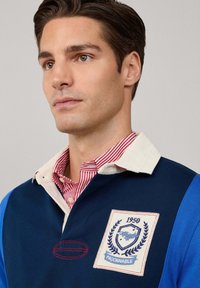 Young man with brown hair wearing a blue and white rugby shirt with a red and white striped collar shirt underneath.