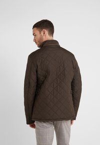 Casaco acolchoado castanho com um colarinho alto, apresentando costura em forma de diamante e ajustes laterais, usado sobre calças xadrez.