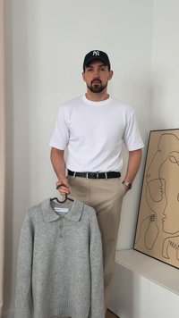 Grå stickad tröja med krage och tre knappar, hängande på en svart galge, bredvid en person som bär en vit t-shirt och beige byxor.