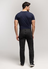 Mężczyzna tyłem, ubrany w granatowy T-shirt, czarne dżinsy o prostych nogawkach i czarne sneakersy na tle jasnego, jednolitego tła.