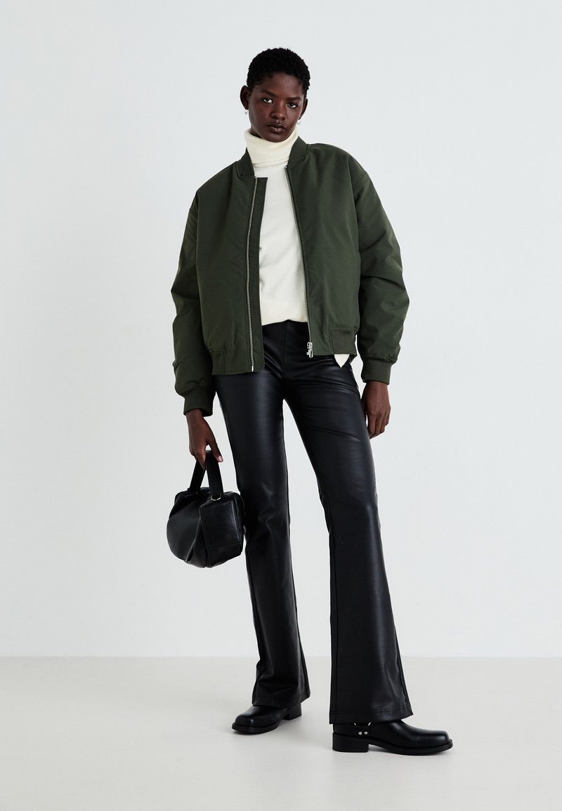 Giubbotto bomber verde, dolcevita bianco, pantaloni in pelle nera a zampa d'elefante, borsa nera e stivali alla caviglia. Texture lisce e design minimalista.