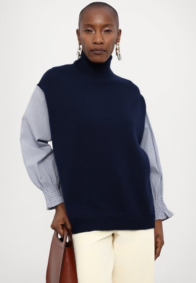 Navy blue turtleneck sweater with gray striped puff sleeves, featuring textured knit at the body and elasticized cuffs. Brown handbag held in hand.
