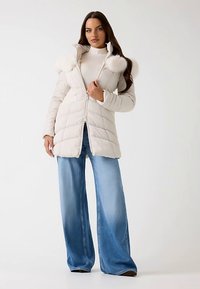 Biała kurtka puchowa z zamkiem z przodu, wyposażona w wysoki kołnierz i ozdoby z sztucznego futra. Połączona z szerokimi, niebieskimi dżinsami o szerokich nogawkach. Gładka faktura.
