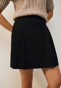 Jupe noire plissée à la texture lisse, avec une taille cintrée et une coupe évasée, associée à un pull en maille marron clair.