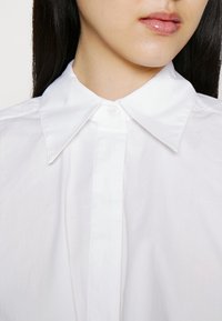 Gros plan d'une personne portant une chemise blanche à col avec boutons, avec de longs cheveux foncés visibles derrière les épaules.
