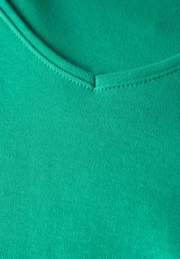 Zelená bavlněná látka s texturovaným pletením, s kulatým výstřihem a plochými švy. Materiál vypadá jemně a hladce.