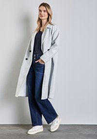 Vaaleanharmaa villakangas takki, jossa on kaulus ja mustat napit, yhdistettynä tummansinisiin leveälahkeisiin farkkuihin ja valkoisiin tennareihin, joissa on beigen sävyisiä yksityiskohtia.