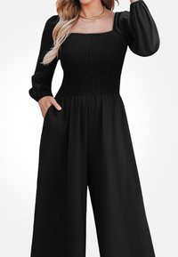 Czarny kombinezon z kwadratowym dekoltem, marszczonym topem, długimi puffowanymi rękawami i szerokimi nogawkami z kieszeniami po bokach. Gładka tekstura materiału.