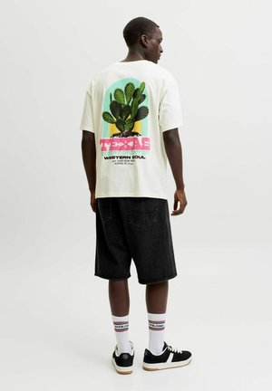 Mand iført hvid T-shirt med kaktus og Texas-tekst på ryggen, sorte knælange shorts, hvide mærkesokker og sorte sneakers står med ryggen til.