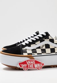 Zapatillas de cuadros en blanco y negro con parte superior de gamuza, cordones blancos y suela de goma con una suela marrón. Incluye una etiqueta roja "Vans Off the Wall".