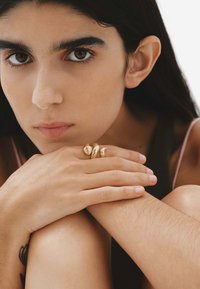 Tres anillos de oro texturizados adornan dedos que descansan sobre piel marrón, con esmalte de uñas natural. El fondo es neutral y suave.