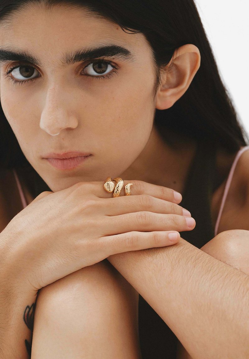 Tres anillos de oro texturizados adornan dedos que descansan sobre piel marrón, con esmalte de uñas natural. El fondo es neutral y suave.