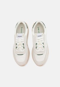 Sneakers Lacoste con tomaia in pelle bianca, accenti in suede beige, dettagli verdi e suole in gomma. Presentano una chiusura con lacci e il logo sulla linguetta.