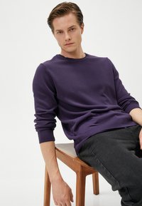 Lila gestrickter Pullover mit Rundhalsausschnitt und gerippten Bündchen, kombiniert mit schwarzen Jeans, Modell sitzt auf einem Holzstuhl vor einem weißen Hintergrund.
