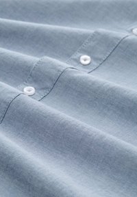 Cămașă din material albastru deschis, cu închideri prin butoane albe și o textură subtilă; material neted, pliat, cu cusături vizibile.