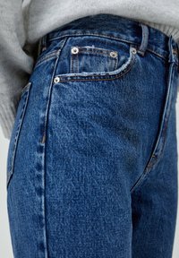 Gros plan d'une personne portant un jean denim bleu avec des coutures orange et un pull gris rentré à la taille.