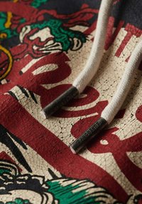 Sudadera con una tela texturizada en crema y un vibrante diseño gráfico agrietado en rojo, verde y negro, y puntas de cordón en gris.