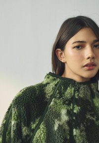 Fleece jakna u nijansama zelene, s visokim ovratnikom, zatvaranjem na patentni zatvarač i teksturiranom, pahuljastom površinom s različitim nijansama.