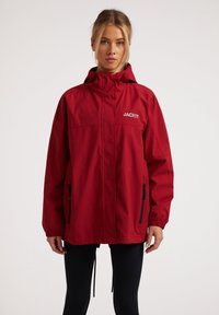 Rød vandtæt jakke med høj krave, forsynet med en frontlynlås, to sorte lynlås-lommer og "JACKIT" logo. Elastiske manchetter og kant.