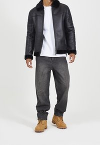 Giacca di pelle nera con colletto e polsini in pelliccia sintetica, t-shirt bianca, jeans grigi a taglio largo e stivali marroni con lacci gialli.