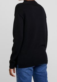 Pull en tricot noir avec une texture côtelée, doté de manches longues et d'évasements sur les côtés, porté sur un jean en denim bleu. Vue arrière affichée.