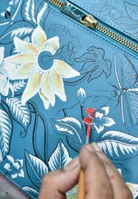 Dekorativer blauer Lederstoff mit weißen Blumenmustern und grünen Blättern. Eine Hand malt mit einem kleinen Pinsel ein rotes Blütendetail in der Nähe eines Reißverschlusses.