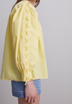 Mujer con blusa amarilla de mangas abullonadas y detalles con cordones, combinada con pantalones cortos de mezclilla remangados, sobre un fondo liso.