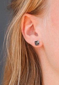 Vierkante zwarte studoorbellen met een facette-oppervlak, gezet in een zilverkleurig metalen frame, getoond op een gedeeltelijk zichtbaar oor.