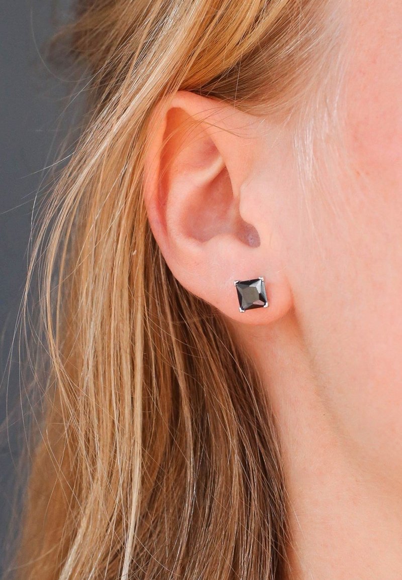 Vierkante zwarte studoorbellen met een facette-oppervlak, gezet in een zilverkleurig metalen frame, getoond op een gedeeltelijk zichtbaar oor.