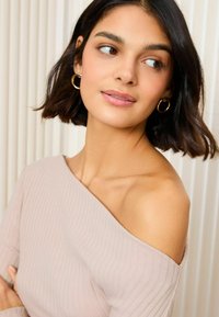 Svetlo béžový off-shoulder rebrovaný top s dlhými rukávmi. Modelka má krátke tmavé vlasy a nosí zlaté kruhové náušnice. Hladký tón pleti, minimálny makeup.