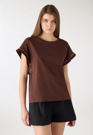 Giovane donna con capelli castani lisci che indossa una maglietta marrone a maniche corte con bordi sfrangiati e pantaloncini neri, in piedi davanti a uno sfondo neutro.