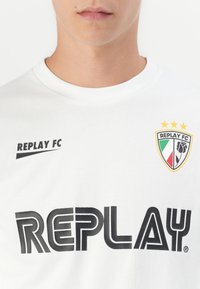 Mand iført hvid t-shirt med sort "REPLAY" tekst, "REPLAY FC" logo og skjoldemblem med tre gule stjerner og blomstermotiv.