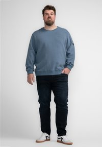 Blauwe katoenen sweatshirt met geribbelde manchetten, gecombineerd met donkerdenim jeans. Model draagt witte sneakers met zwarte accenten. Eenvoudig ontwerp, ontspannen pasvorm.
