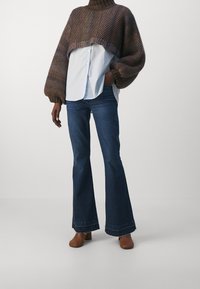 Maglione marrone in stile uncinetto con un collo alto sopra una camicia button-up azzurra, abbinato a jeans svasati blu scuro e scarpe marroni.