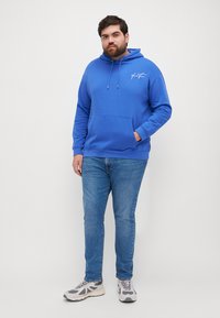 Felpa blu con tasca frontale, cappuccio regolabile con cordino e logo bianco. Indossata con jeans azzurri e scarpe da ginnastica grigie, in piedi su uno sfondo bianco.