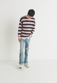 Joven con cabello negro que lleva un suéter a rayas rosa y negro, jeans de color azul claro y zapatillas blancas, mirando hacia abajo sobre un fondo blanco liso.