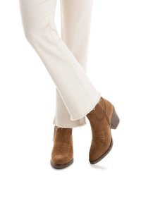 Stivaletti in camoscio marrone con tacco a blocco, caratterizzati da motivi di cucitura decorativi. Indossati con pantaloni bianchi sfilacciati.