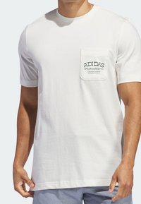 Bílé bavlněné tričko s kulatým výstřihem a krátkým rukávem, které má na levé straně hrudi kapsu s textem "Adidas Groundkeeping" ve zelené barvě.