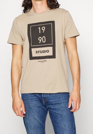 Man wearing beige T-shirt with black square graphic reading "1990 Studio" and blue jeans, standing against plain background.