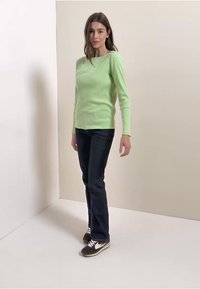 Vaaleanvihreä pitkähihainen raidallinen paita yhdistettynä tummansinisiin farkkuihin ja tummiin tennareihin, seisomassa neutraalia taustaa vasten.