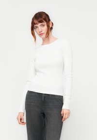 White long-sleeve ribbed shirt with a rounded neckline, paired with dark grey jeans. The fabric appears soft with a fitted design.